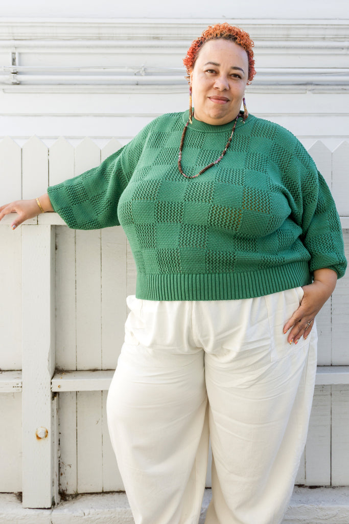 Model wearing Anya Checkerboard Sweater in Fern and Cadence Pants in White, standing in front of white building