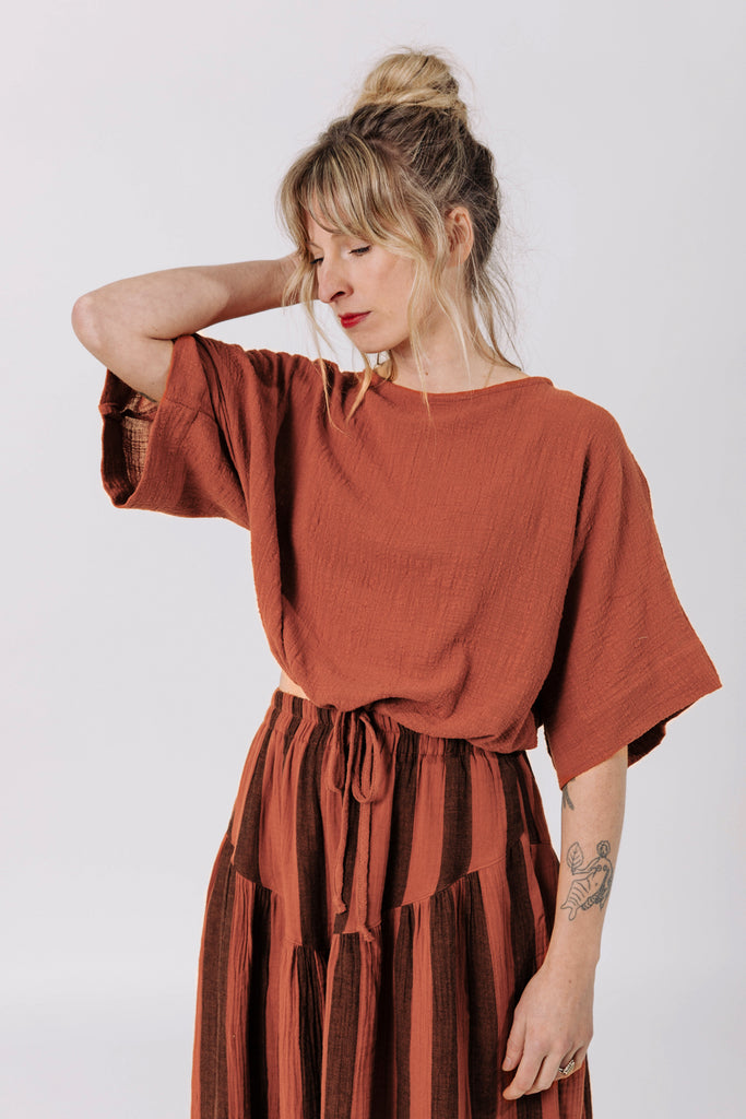 Front view of model wearing Ayana Top in Terracotta and Jessie Skirt in Terracotta Stripe, standing with one hand behind neck in front of white background