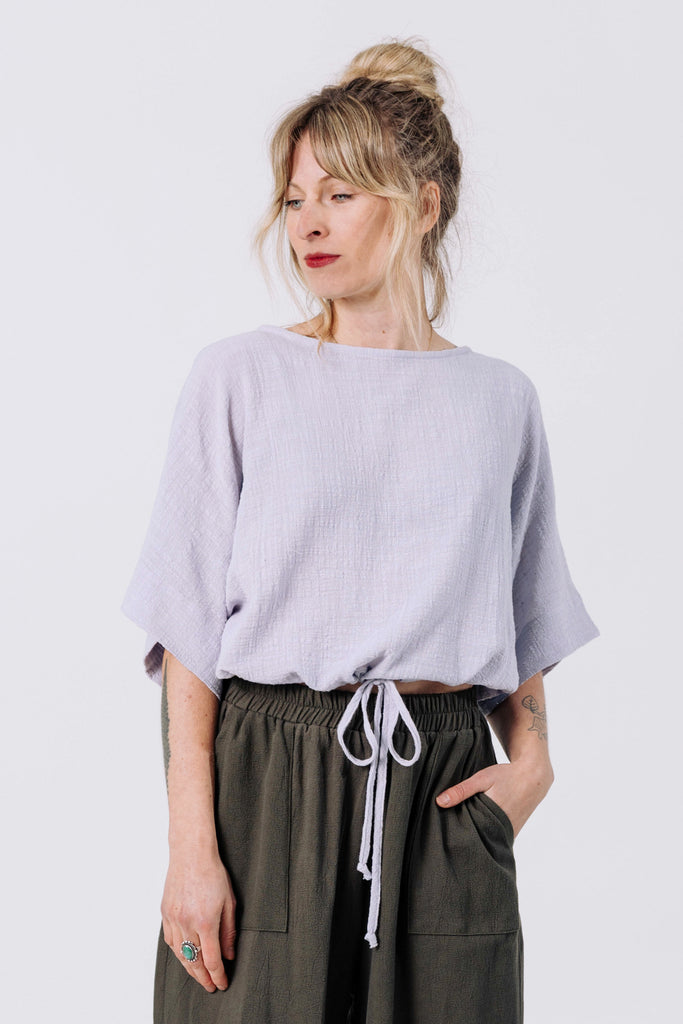 Front view of model wearing Ayana Top in Mist and Atlas Pant in Mud, standing with one hand in pant pocket in front of white wall