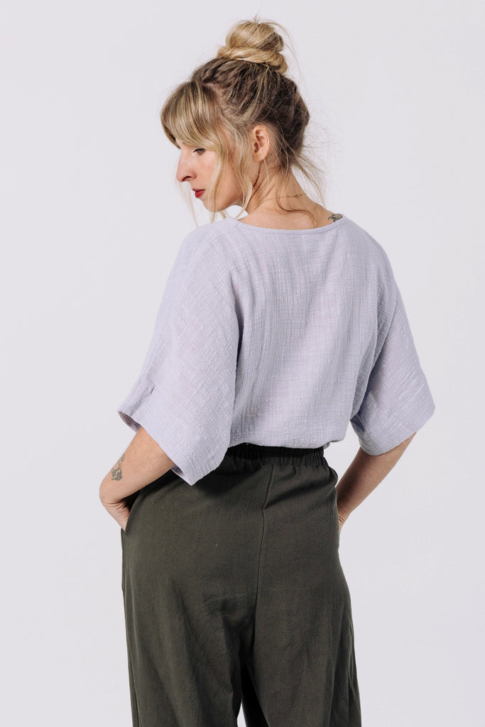 Back view of model wearing Ayana Top in Mist and Atlas Pant in Mud, standing with hands in pant pockets in front of white background
