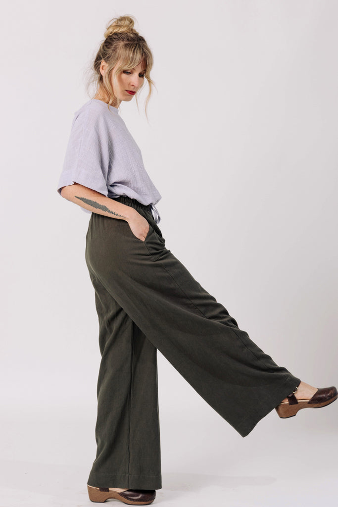 Side view of model wearing Ayana Top in Mis, and Atlas Pant in Mud, standing with hands in pant pockets in front of white wall