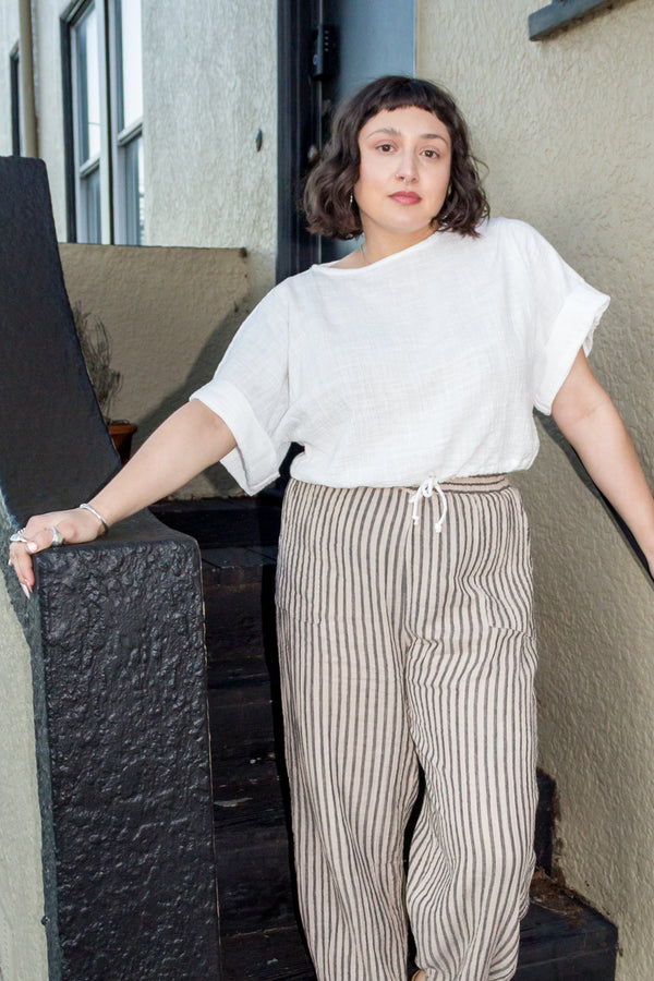 Model wearing Ayana Top in White and Cadence Pant in Oat Stripe, standing with in black and tan stair entryway