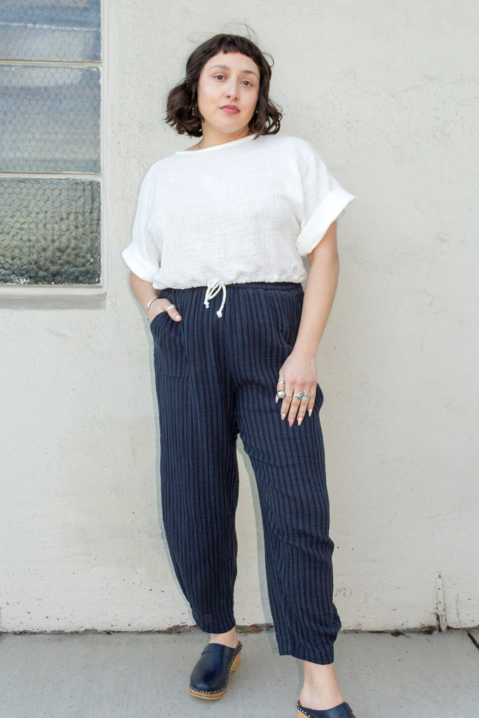 Model wearing Ayana Top in White and Cadence Pant in Black Stripe, standing with one hand in pant pocket in front of white wall