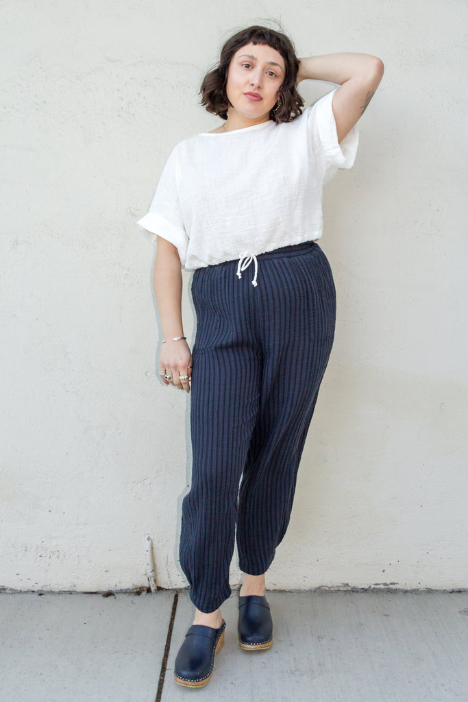 Model wearing Ayana Top in White and Cadence Pant in Black Stripe, standing with one hand on top of head in front of white wall