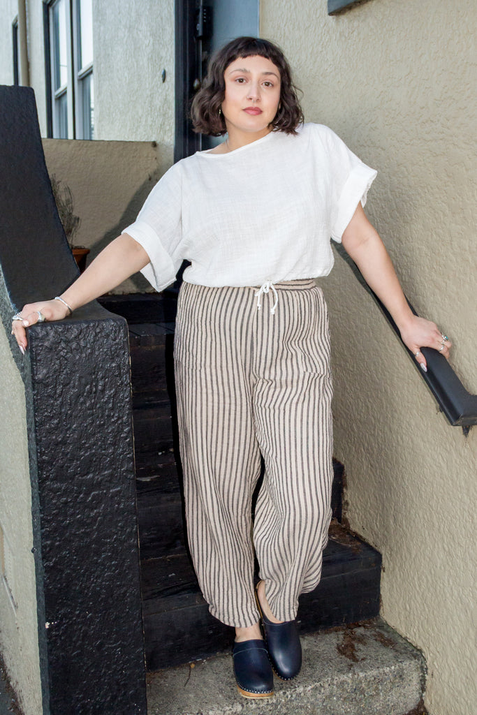 Model wearing Ayana Top in White and Cadence Pant in Oat Stripe, standing with in black and tan stair entryway
