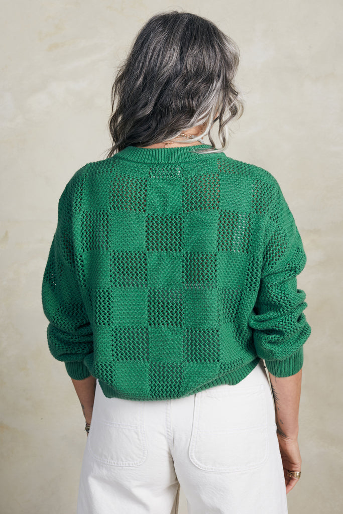 Back view of model wearing Anya Checkerboard Sweater In Fern and white jeans in front of white background