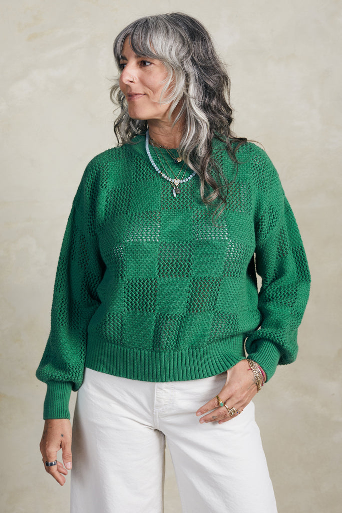 Model wearing Anya Checkerboard Sweater In Fern and white jeans in front of white background