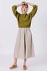 Model wears Clove Sweater in Fennel and Fawn Poplin Skirt in Oatmeal, standing with hands above their head, in front of white background