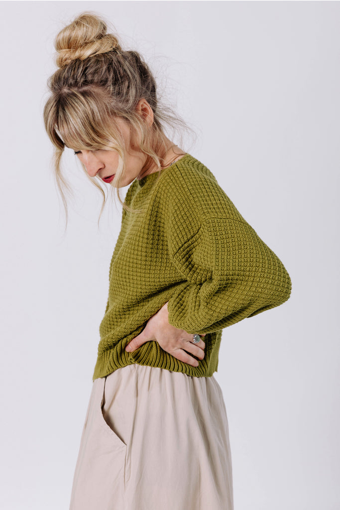 Side view of model with arms over head wearing Clove Sweater in Fennel and Fawn Skirt in Oat in front of white background
