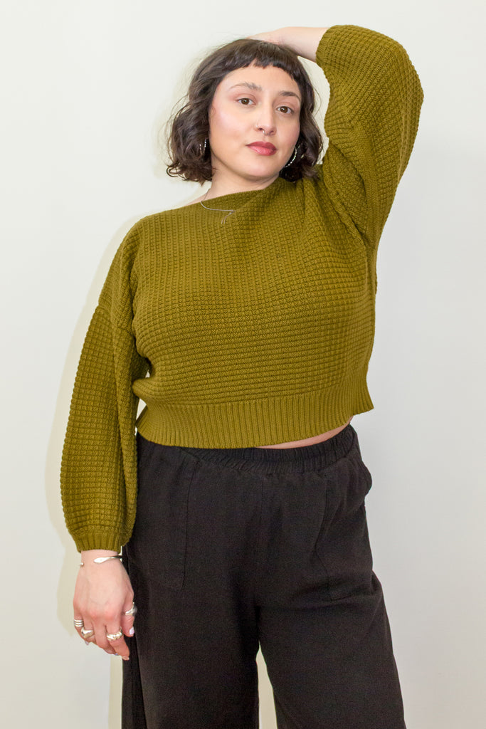 Model wearing Clove Sweater in Fennel and wide leg pants in black with hand raised to head, standing in front of white background
