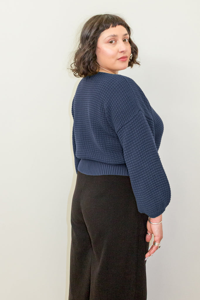 Back/side view of model wearing Clove Sweater in Fennel and wide leg pants in black standing in front of white background