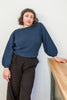 Model wearing Clove Sweater in Indigo and wide leg pants in black leaning on table in front of white background