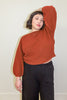 Model wearing Clove Sweater in Paprika and wide leg pants in black with hand raised to head, standing in front of white background