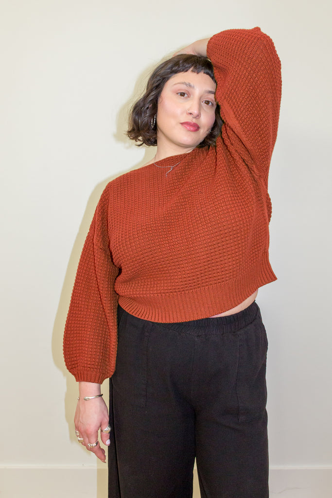 Model wearing Clove Sweater in Paprika and wide leg pants in black with hand raised to head, standing in front of white background