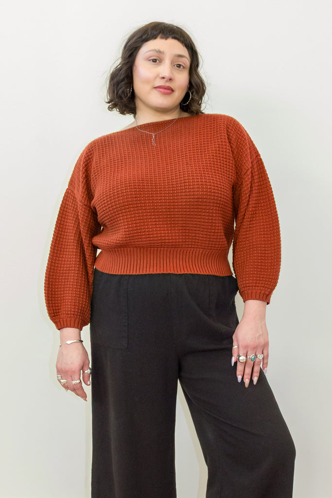 Model wearing Clove Sweater in Paprika and wide leg pants in black standing in front of white background