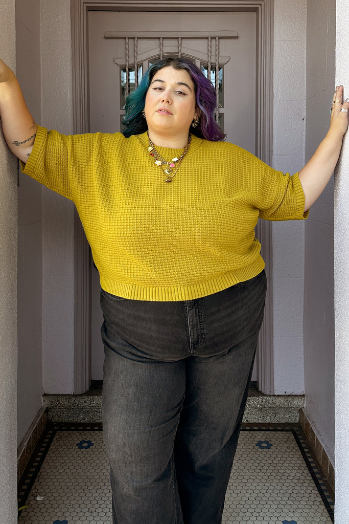 Person wearing a dijon Cody sweater and dark pants standing in a hallway.