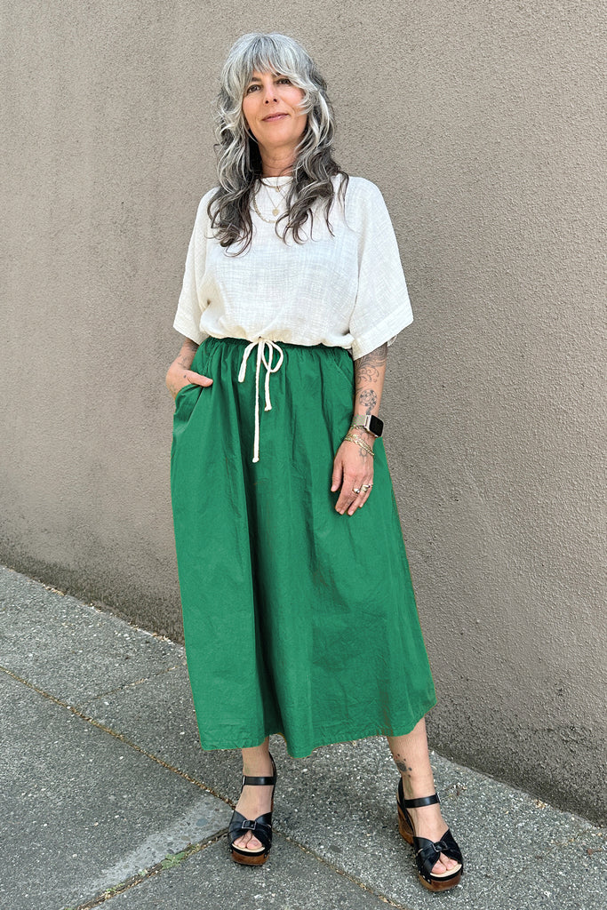 Woman wearing a white ayana top and fern green Fawn Skirt against a beige wall.