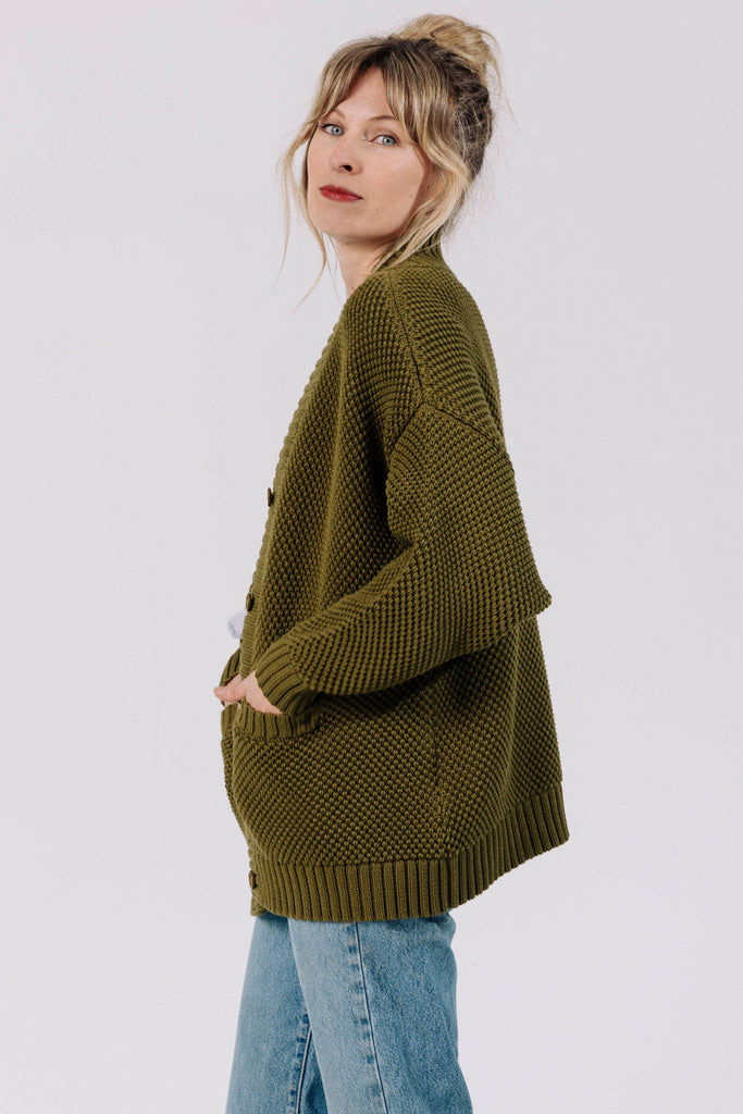 Side view of model wearing Frankie Cardigan in Fennel and light wash jeans with hands in cardigan pockets, in front of white background