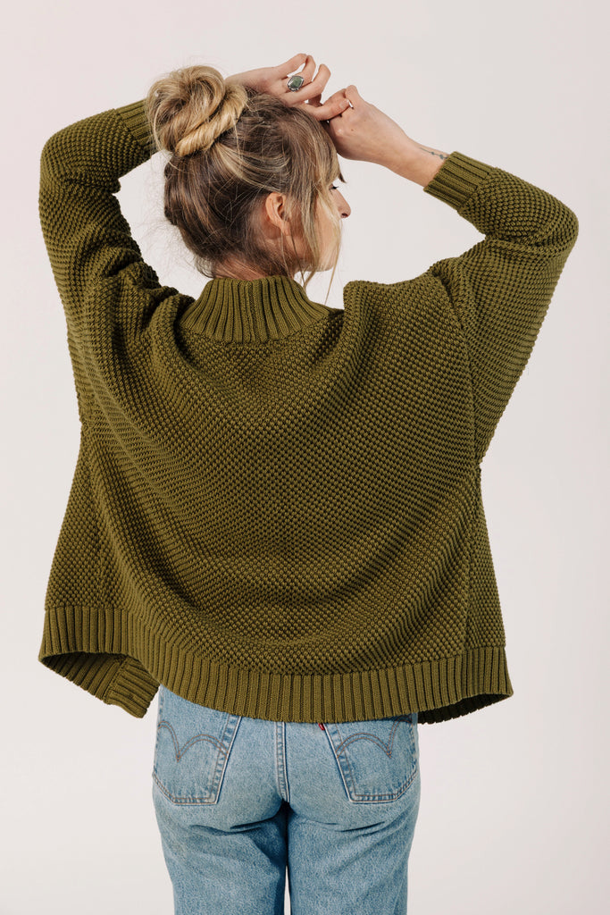 Back view of model wearing Frankie Cardigan in Fennel and light wash jeans, raising arms over head in front of white background