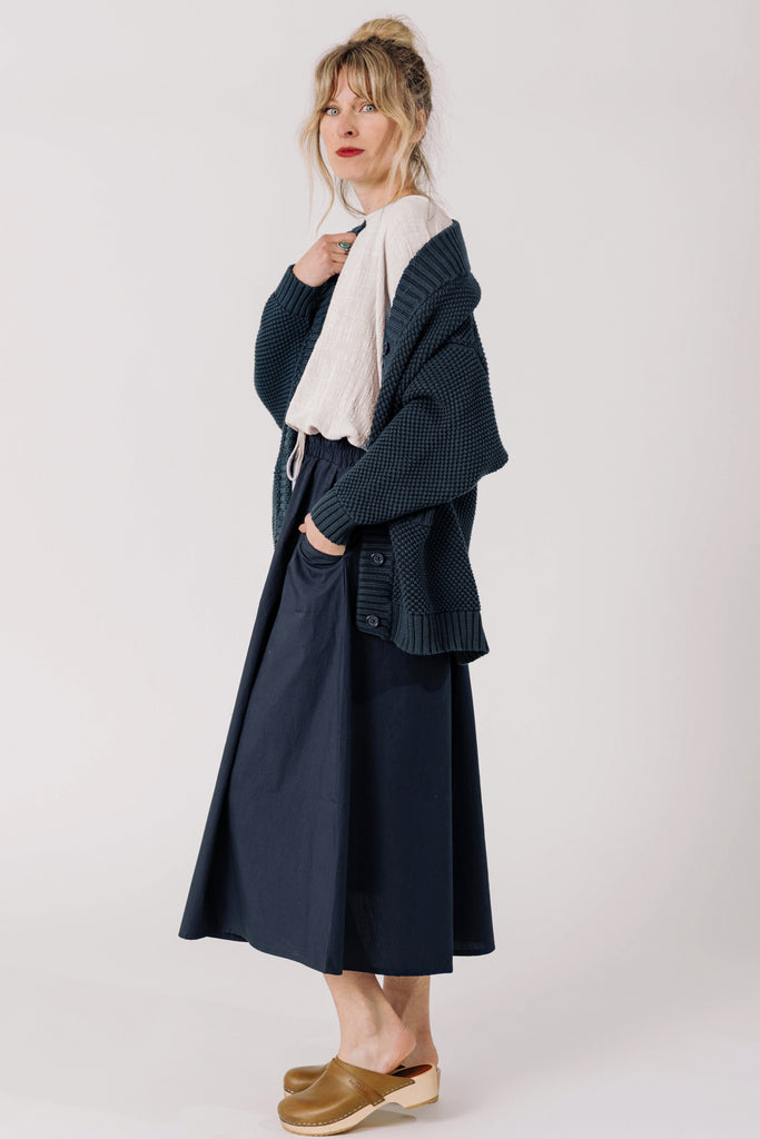 Side view of model wearing Lino Cardigan in Indigo, Ayana Top in Oyster, and Fawn Poplin Skirt in Indigo in front of white background