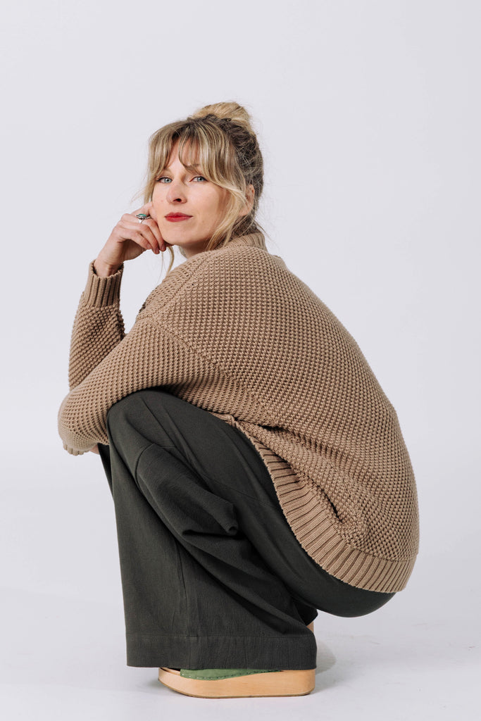 Side view of model wearing Frankie Cardigan in Tan and Atlas Pant in Mud, squatting in front of white background