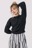 Front view of model wearing Ivy Sweater in Black and Jessie Stripe Skirt in Oyster, with one hand raised to back of the head, standing in front of white background
