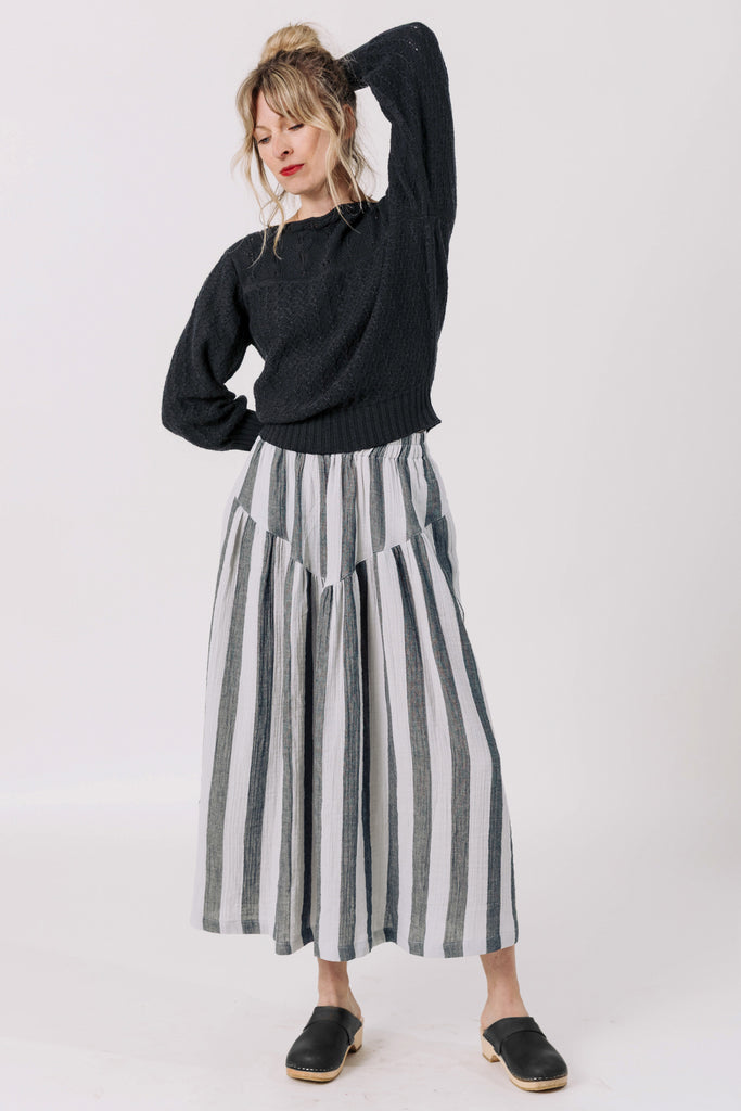 Front view of model wearing Ivy Sweater in Black and Jessie Stripe Skirt in Oyster, with one hand raised to back of the head, standing in front of white background