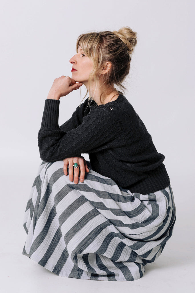 Side view of model wearing Ivy Sweater in Black and Jessie Stripe Skirt in Oyster, with one hand under chin and one resting across knees, squatting in front of white background
