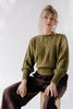 Model wearing Ivy Sweater in Fennel and Atlas Pant in Chocolate, sitting on wooden box stool in front of white background