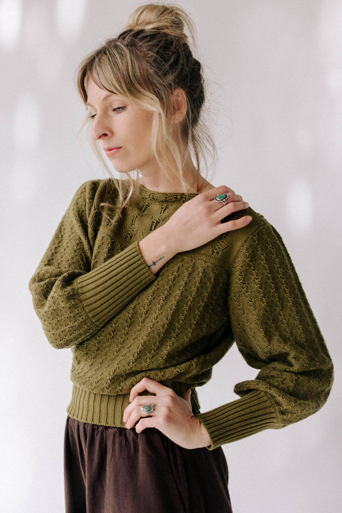 Model wearing Ivy Sweater in Fennel and Atlas Pant in Chocolate standing with one hand on hip and one on shoulder in front of white background