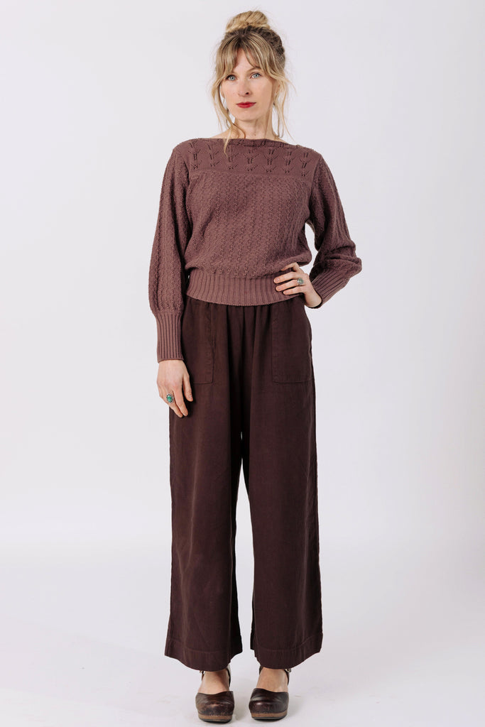 Front view of model wearing Ivy Sweater in Raisin and Atlas Pant in Chocolate, standing with one hand on hip in front of white wall