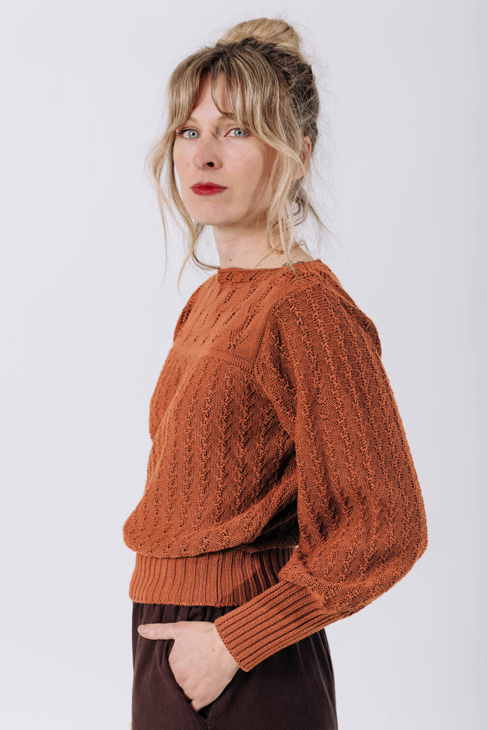 Side view of model wearing Ivy Sweater in Ivy and Atlas Pant in Chocolate, standing with one hand in pocket in front of white wall