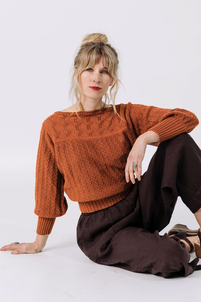 Front view of model wearing Ivy Sweater in Rust and Atlas Pant in Chocolate, sitting with one elbow rested on raised knee in front of white wall