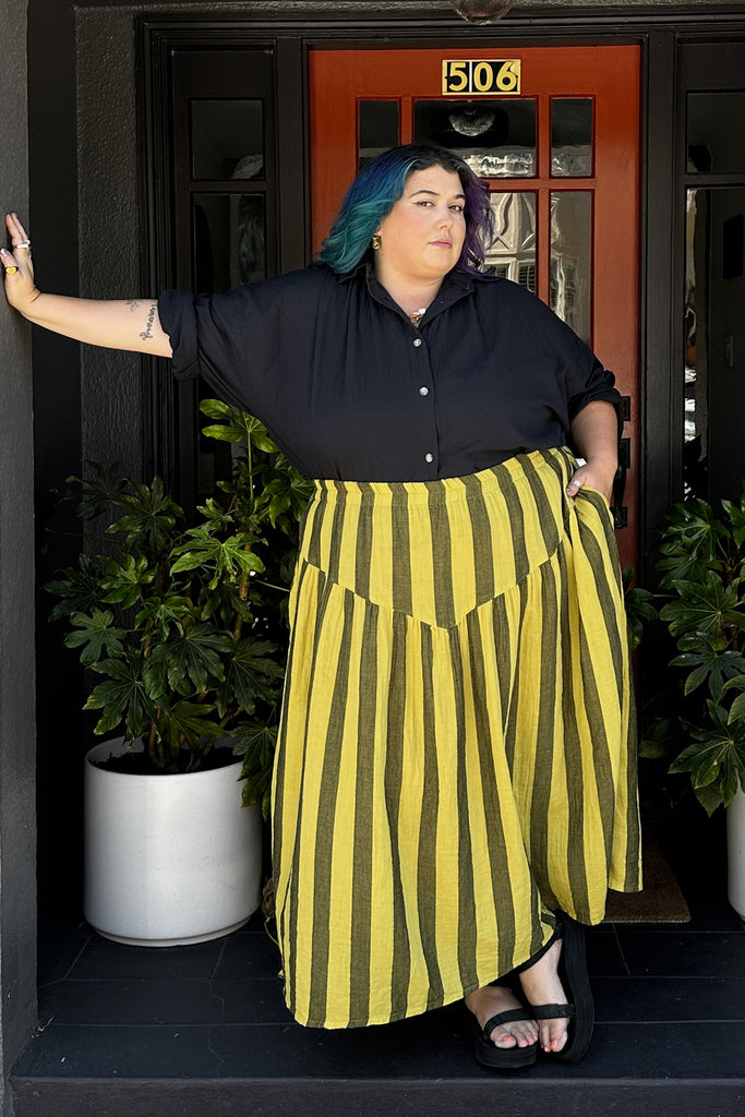Person wearing a black poet shirt and dijon striped jessie skirt standing in front of a door with a number 506.