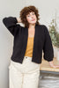 Model wearing Jonah Cardigan in Black and Painter Pant in Natural leaning on wooden tabletop in front of white wall