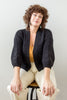 Model wearing Jonah Cardigan in Black and Painter Pant in Natural sitting on wood and black metal stool in front of white wall