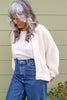 Model wearing Boucle Jonah Sweater in Natural, Minnie Tank in White, and blue jeans, standing in front of green wall