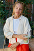 Model wearing Boucle Jonah Cardigan in Natural, Maisie Longsleeve in Oyster, and orange bottoms standing in front of black gate with green vines 