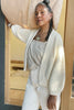 Model wearing Boucle Jonah Cardigan in Natural, Bianca Top in Oat, and white jeans, standing in front of wooden doorway of grey building