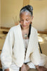 Model wearing Boucle Jonah Cardigan in Natural, Bianca Top in Oat, and white jeans, sitting in front of light yellow wall