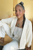 Model wearing Boucle Jonah Cardigan in Natural, Bianca Top in Oat, and white jeans, sitting in front of light yellow wall