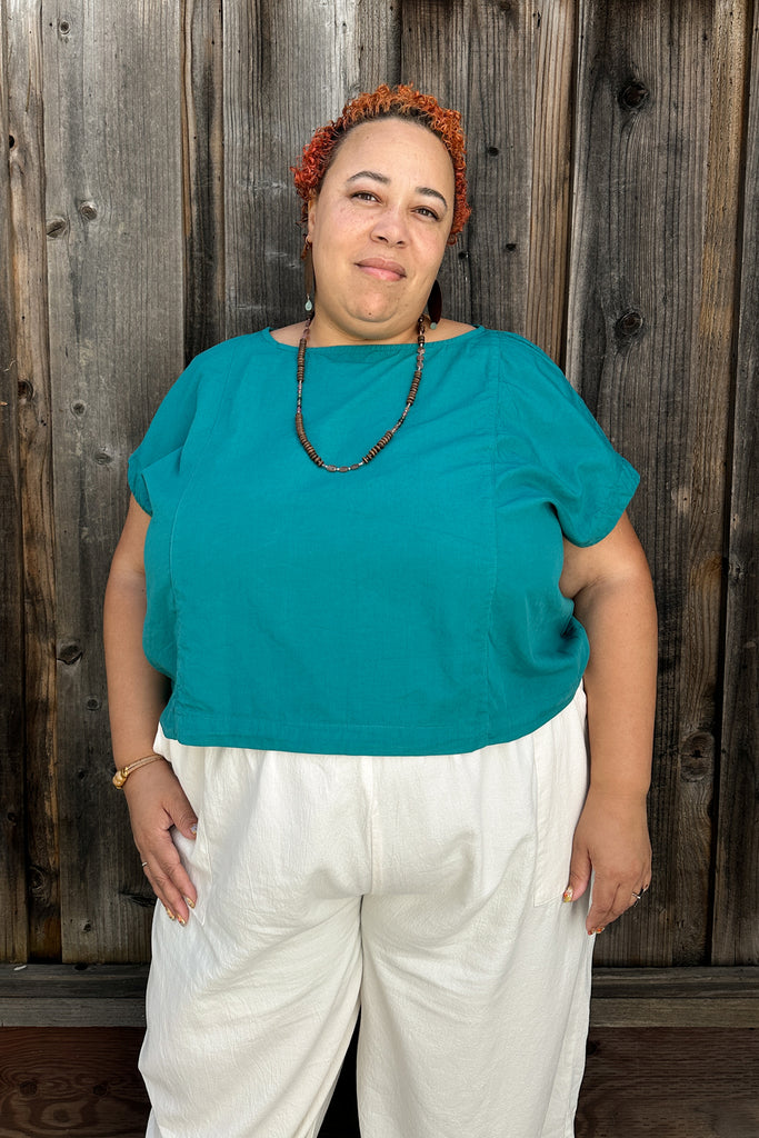 Model wearing Jones Top in Ocean and Cadence Pants in White, standing in front of a dark wooden background