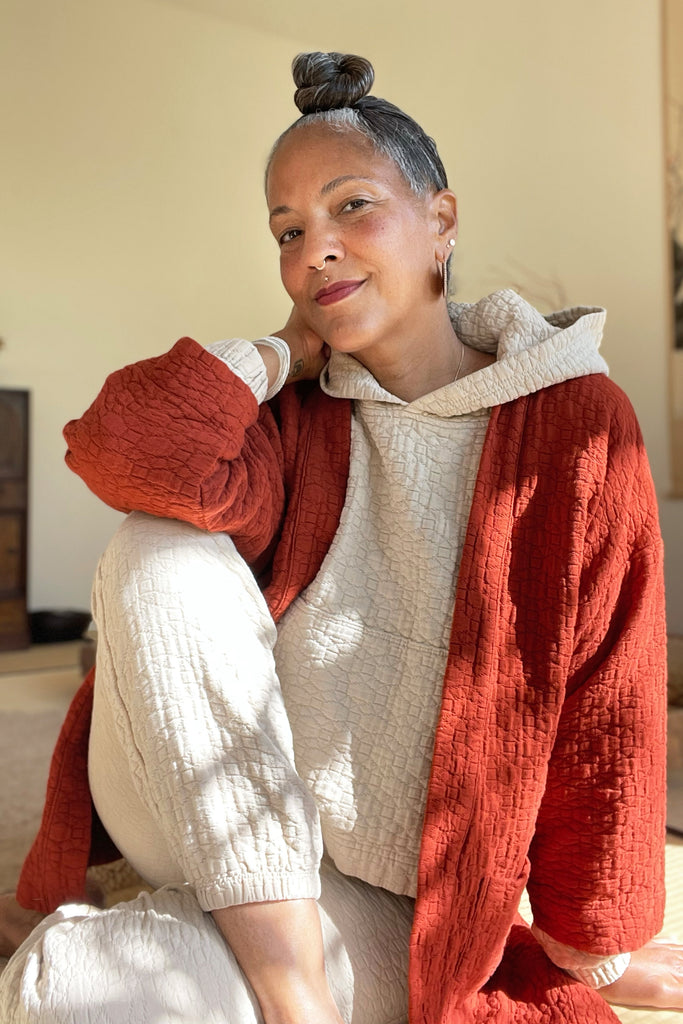 Front view of model wearing Jules Cardigan in Brick, Quilty Hoodie in Oat, and Quilty Joggers in Oat sitting in on matted floor