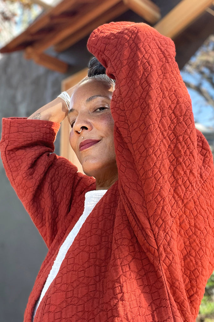 Side view of model wearing Jules Cardigan in Brick with hands raised to head in front of grey building