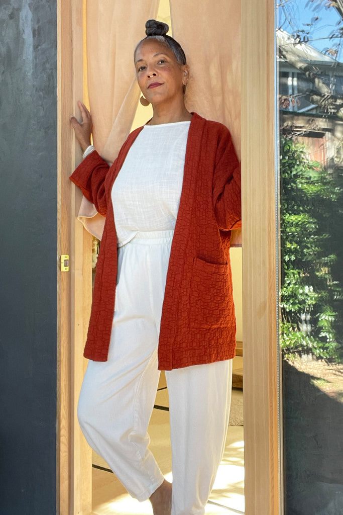 Side view of model wearing Jules Cardigan in Brick, Ayana Top in White, and Cadence Pant in Natural, standing in wooden doorway