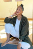 Front view of model wearing Jules Cardigan in Mud, Ayana Top in White, and Cadence Pant in Natural, sitting on wooden floor with one hand raised to back of neck