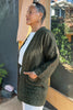 Side view of model wearing Jules Cardigan in Mud, Ayana Top in White, and Cadence Pant in Natural, standing with one hand in cardigan pocket in front of grey building with glass window