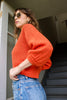 Side view of model wearing Lana Sweater in Tomato and light wash jeans, standing in open doorway with stairs leading into the building