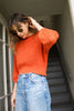 Model wearing Lana Sweater in Tomato and light wash jeans, standing in open doorway with stairs leading into the buildiing