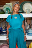 Model wears Jones Top in Ocean and Larkin Pant in Ocean, standing in front of shelves holding colorful bowls and plates.